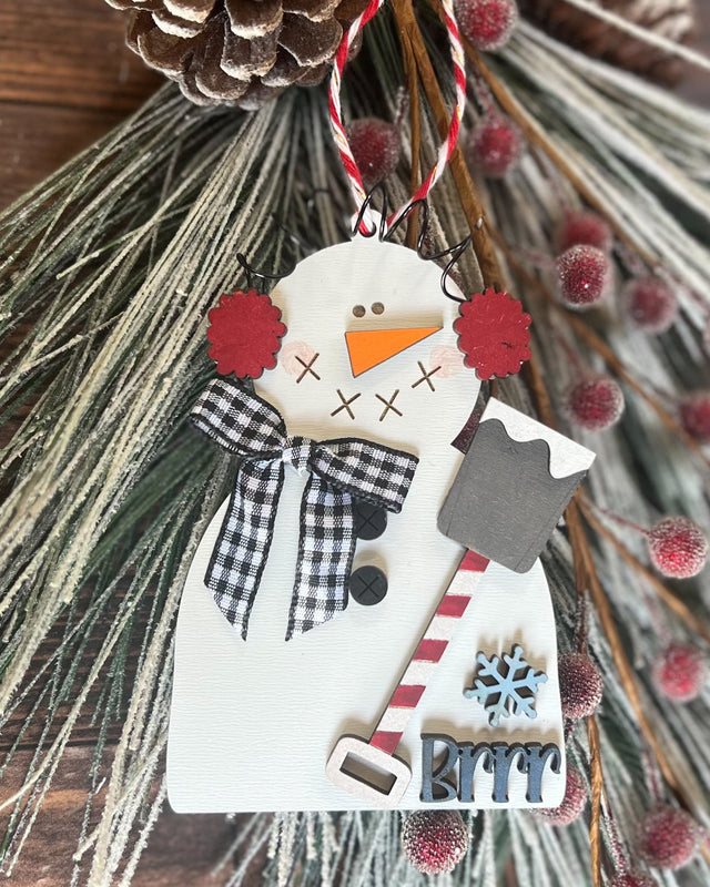 A wooden snowman ornament with a black and white gingham scarf, red earmuffs, and a carrot nose. It has a 'Brrr' lettering and a blue snowflake design.