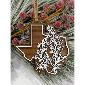 Texas-shaped wooden ornament with white floral overlay and twine hanger displayed on frosted greenery with red berries.