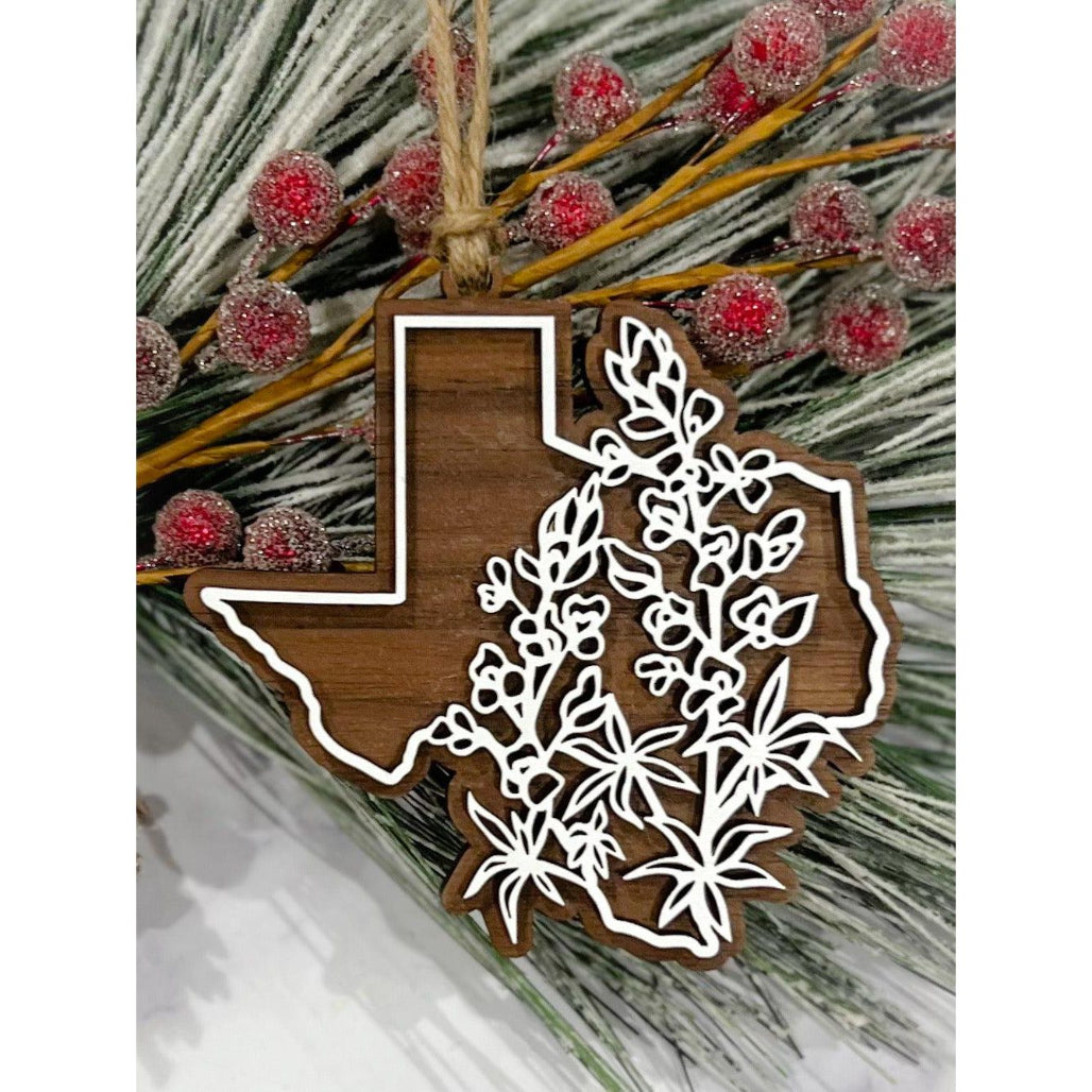 Texas-shaped wooden ornament with white floral overlay and twine hanger displayed on frosted greenery with red berries.
