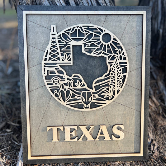 A laser-cut wooden sign or ornament with a silhouette of Texas and iconic symbols, including an oil rig, desert cacti, and the Texas sun. The word 'TEXAS' is prominently displayed at the bottom.