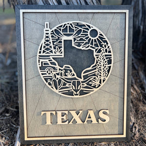 A laser-cut wooden sign or ornament with a silhouette of Texas and iconic symbols, including an oil rig, desert cacti, and the Texas sun. The word 'TEXAS' is prominently displayed at the bottom.