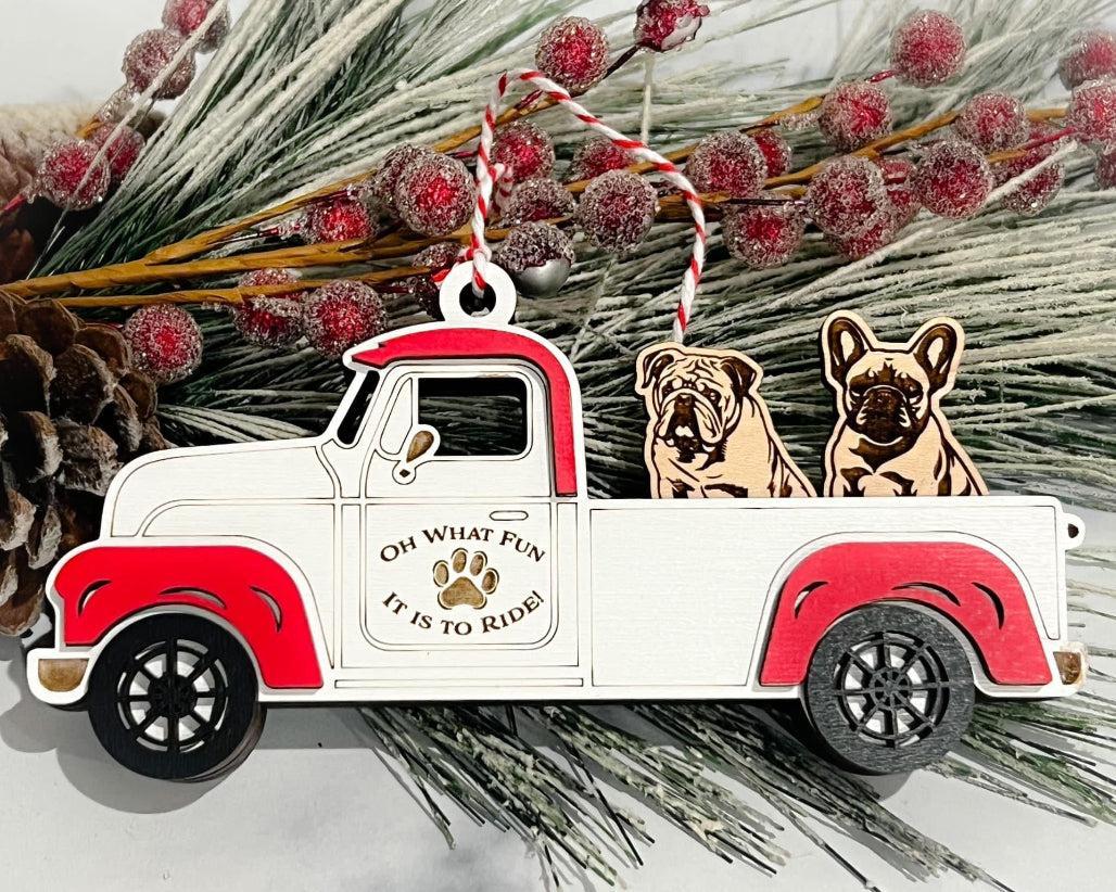 A vintage style white and red truck ornament with two dog figures inside, set against a backdrop of pine cones and decorative branches.
