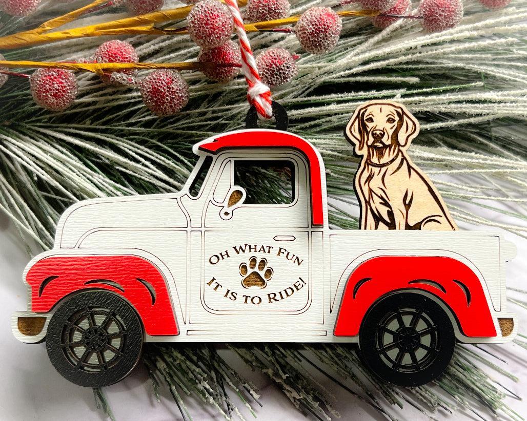 A vintage style red and white truck ornament with a dog riding in the back seat, placed on a branch with Christmas decorations.