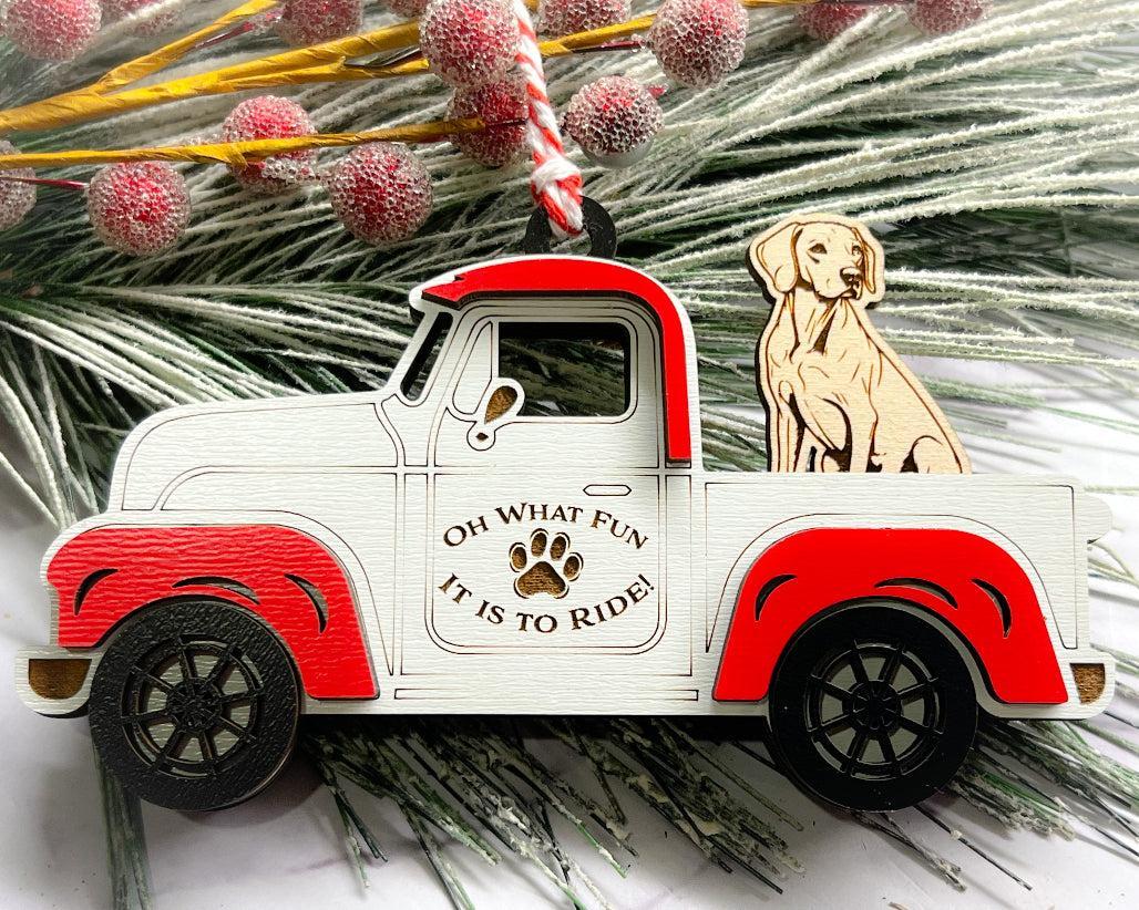 A vintage style red and white truck ornament with a dog riding in the back seat, placed on a branch with Christmas decorations.