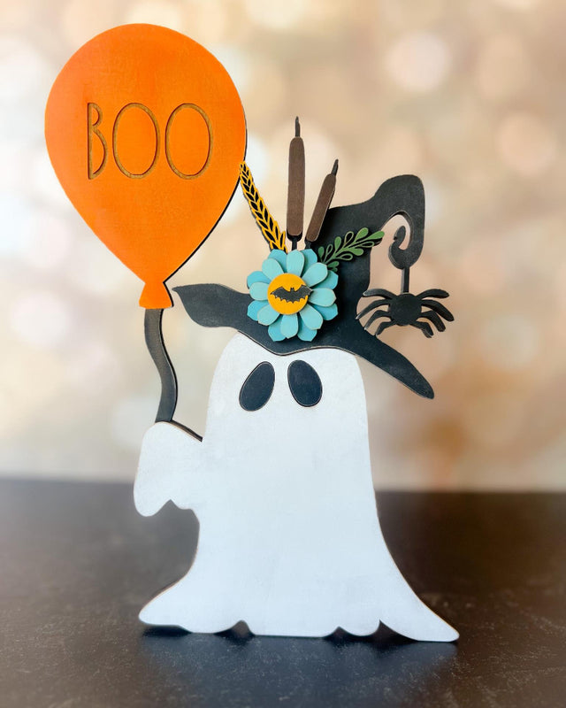 Decorative ghost figure with a witch hat, flower, and orange 'BOO' balloon on a blurred background.