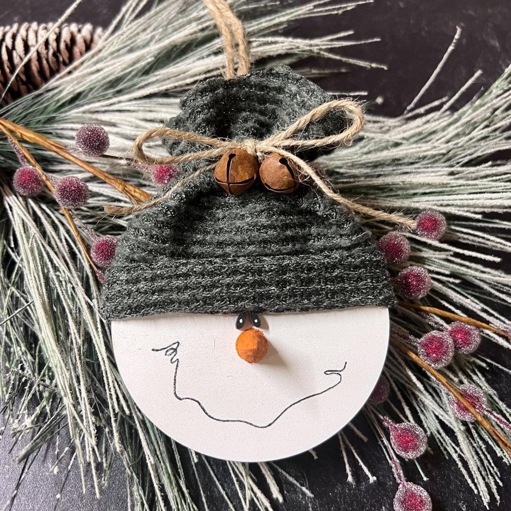 Decorative snowman ornament with a gray hat and orange nose, surrounded by greenery and berries.