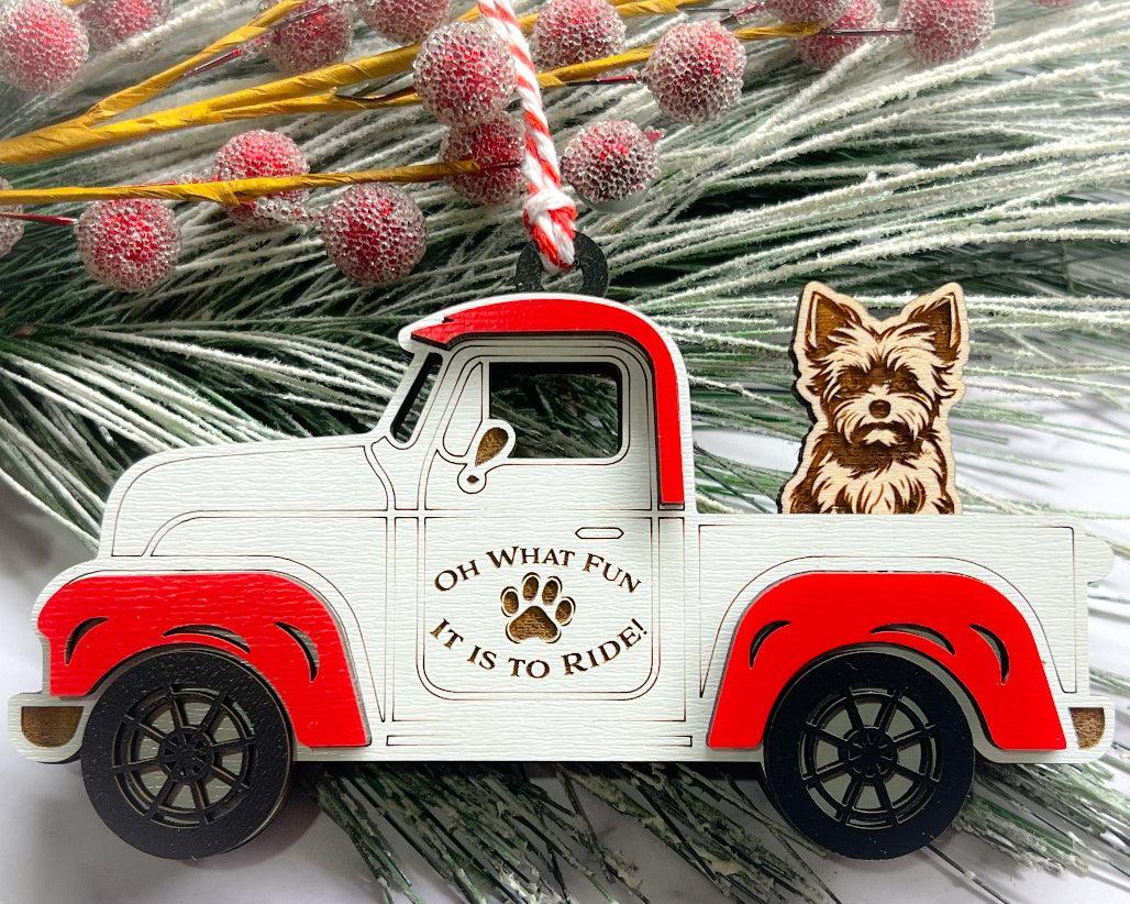 A vintage style red and white truck ornament with a dog riding in the back seat, placed on a branch with Christmas decorations.