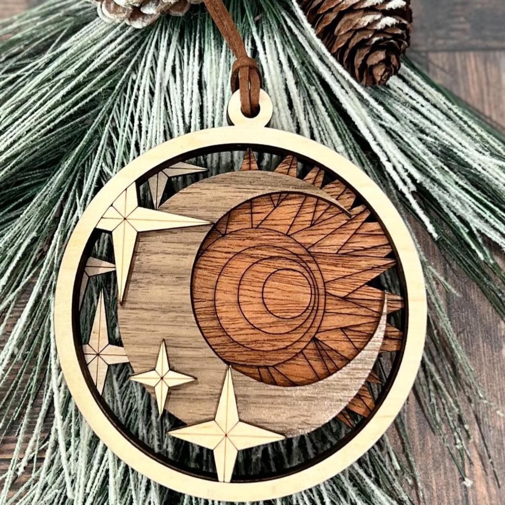 A circular wooden ornament with a laser-cut design featuring the sun, moon, and stars, hanging from a pine branch.
