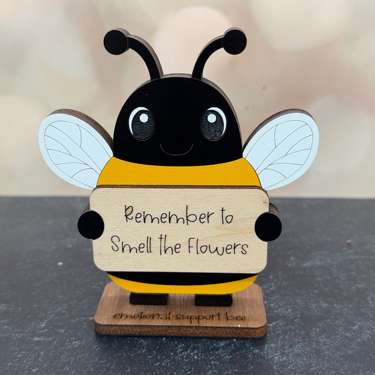 A wooden bee-shaped decorative sign with a black and yellow body, white wings, and a natural wood sign that reads 'Remember to Smell the Flowers.' It stands on a small wooden base against a softly blurred beige background.