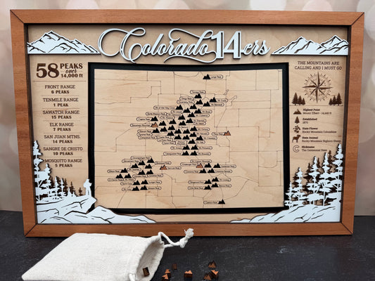 A framed wooden Colorado 14ers map plaque with engraved mountain scenery, a detailed trail map, and informational text, displayed on a dark tabletop with a small drawstring bag and wooden markers in front.