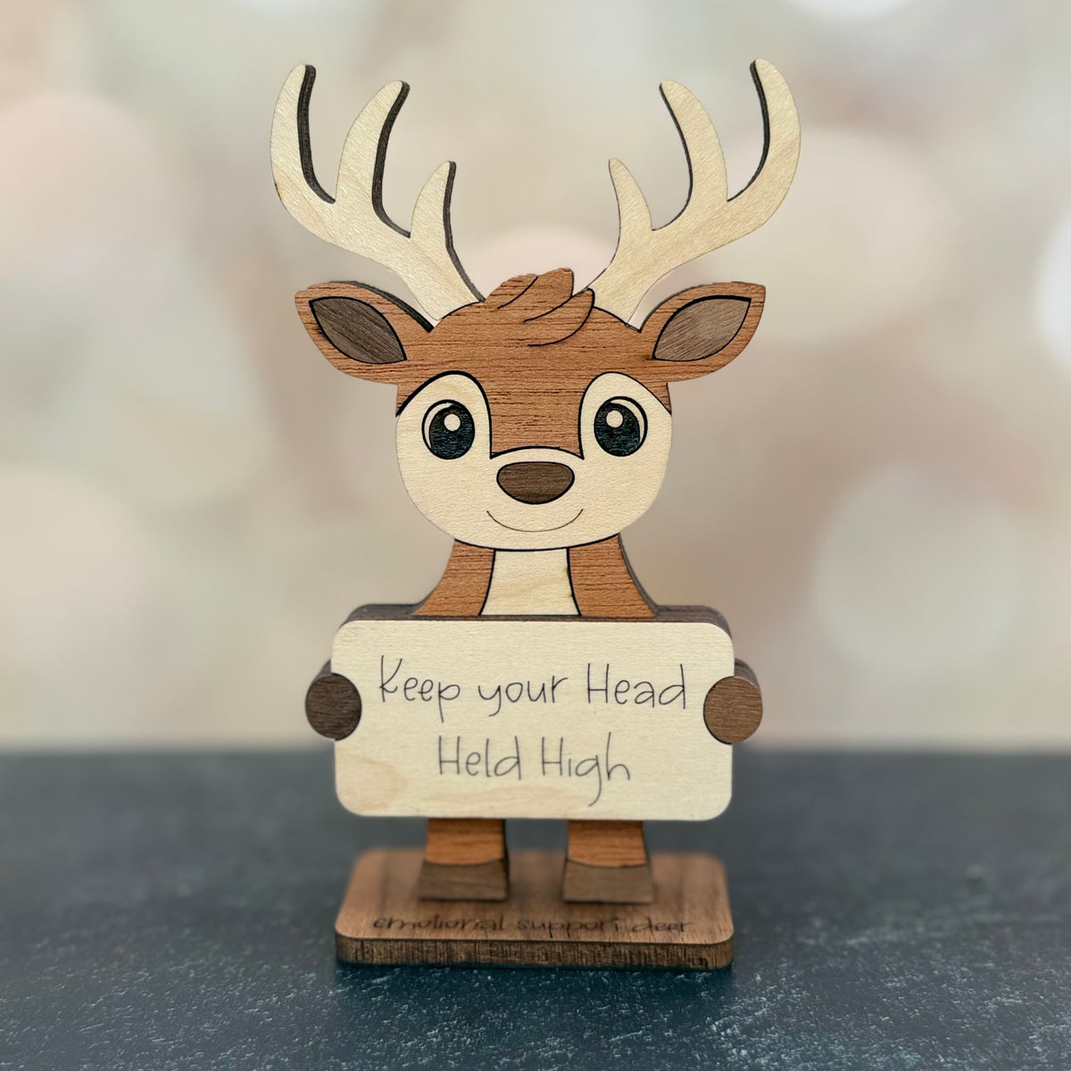 A small wooden deer figurine with antlers holding a sign that reads 'Keep your Head Held High,' photographed on a dark surface with a softly blurred warm-toned background.