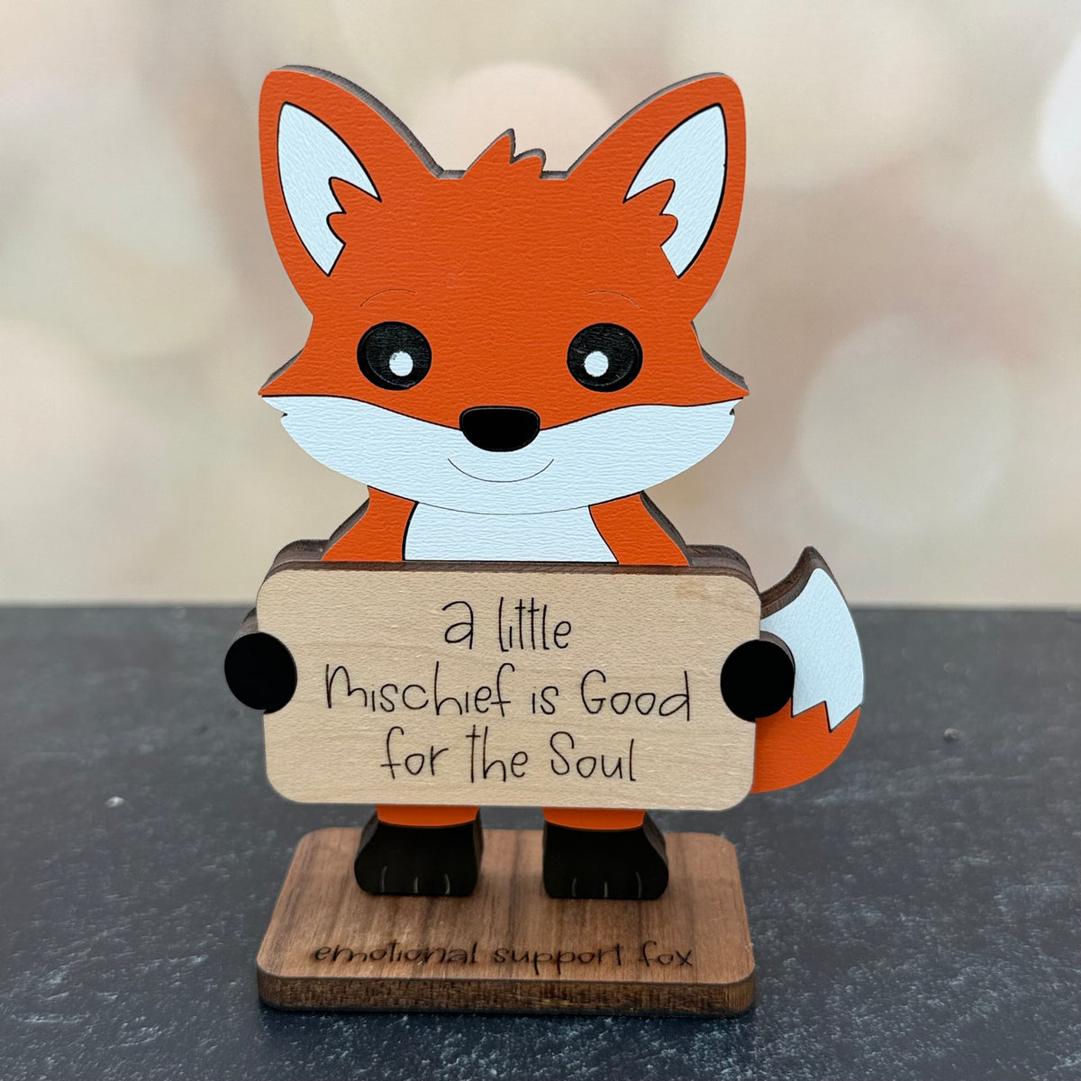 A wooden fox figurine holding a sign that reads 'a little Mischief is Good for the Soul', displayed on a dark gray surface with a soft-focus beige background.