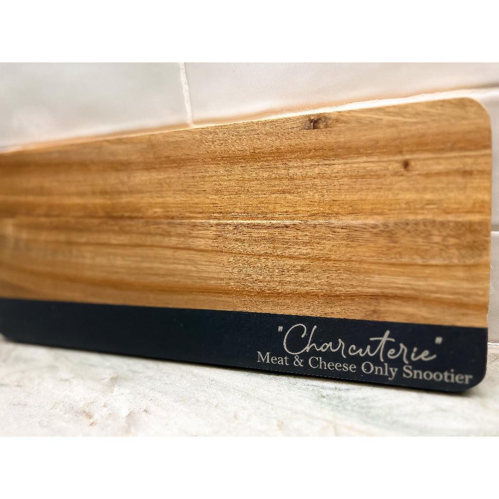 Wooden charcuterie board with engraved phrase “Charcuterie Meat & Cheese Only Snootier,” displayed on a marble countertop.