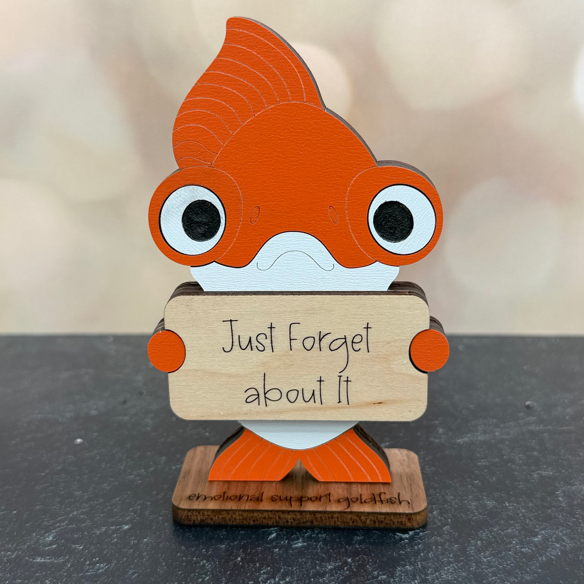 A wooden goldfish-shaped sign holding a small wooden plaque that reads 'Just Forget about It.' The goldfish is orange and white, mounted on a dark surface against a softly blurred beige background.