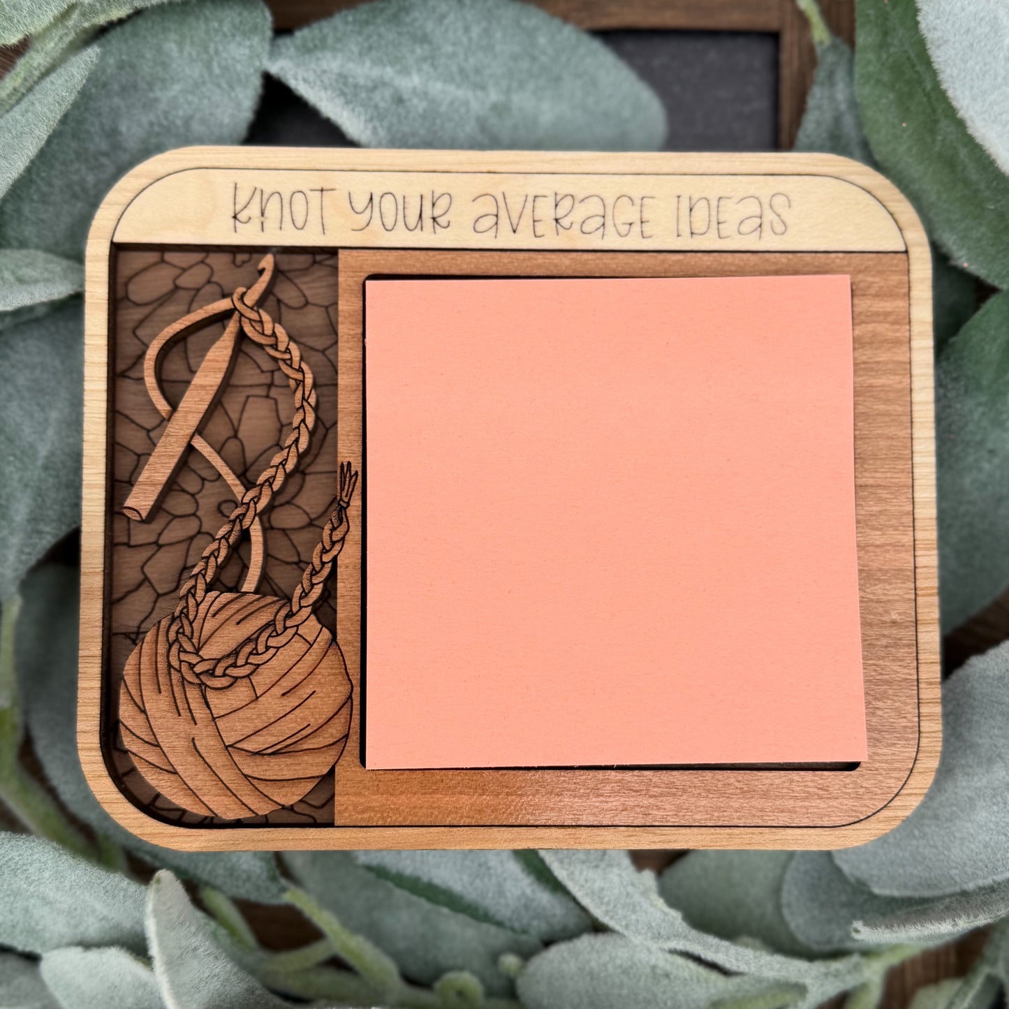 Wooden coaster with a ball of yarn design and a pink square on a green leaf background