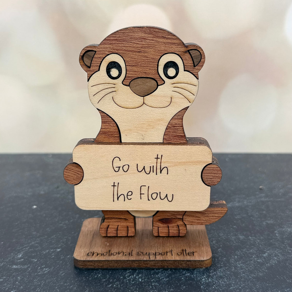 A wooden otter-shaped emotional support sign holding a card that reads 'Go with the Flow', standing on a base engraved with 'emotional support otter'.