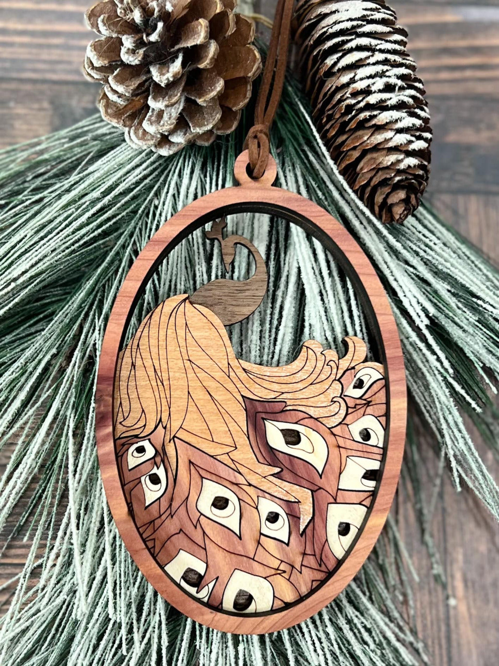 A decorative peacock ornament with intricate details, hanging from a greenery wreath, against a wooden background with pine cones.