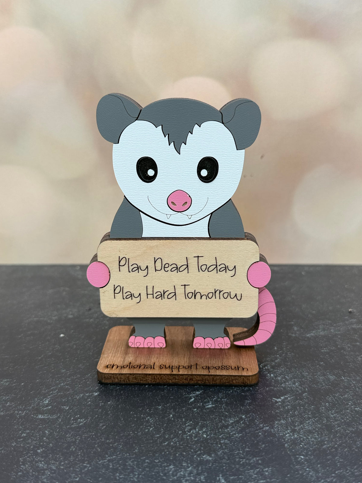 A cute possum-shaped desk sign holding a wooden sign that reads 'Play Dead Today Play Hard Tomorrow' on a dark gray surface with a blurred beige bokeh background.