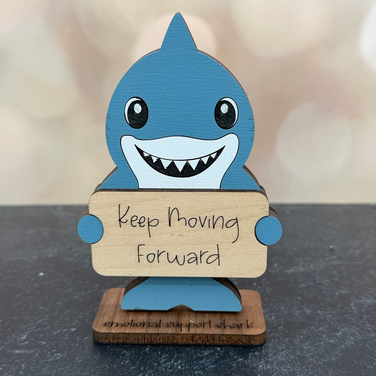 A handcrafted wooden shark figurine painted in shades of blue and white, holding a wooden sign that reads 'Keep Moving Forward'. The base is engraved with 'emotional support shark' and sits on a dark gray surface with a soft-focus beige background.