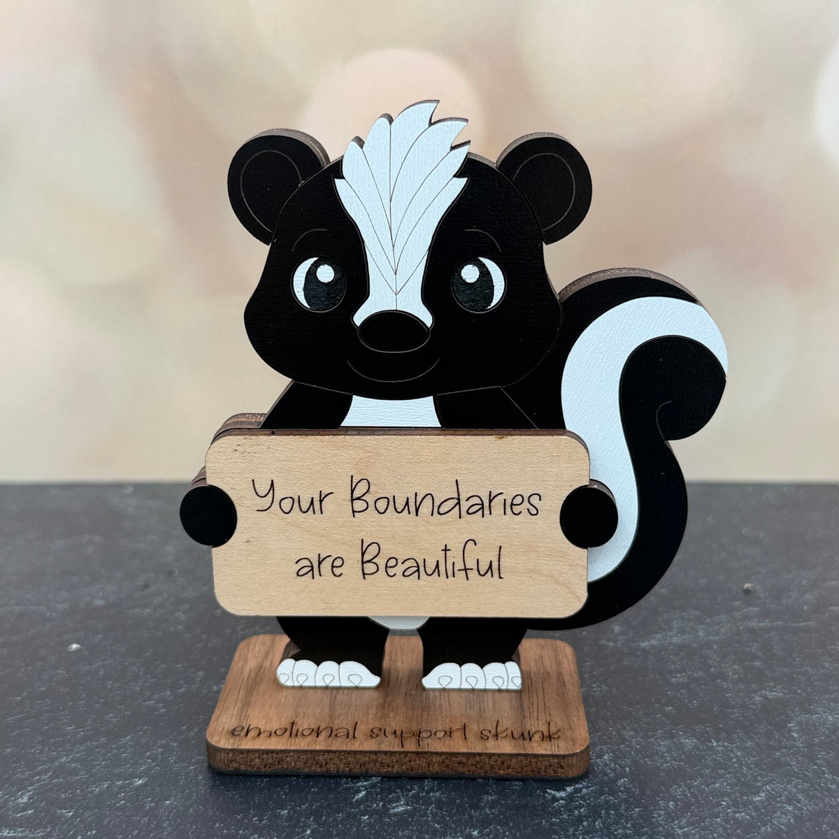 A wooden skunk-shaped sign holder with the engraved message 'Your Boundaries are Beautiful' on its front, standing on a dark gray surface with a soft-focus beige background.