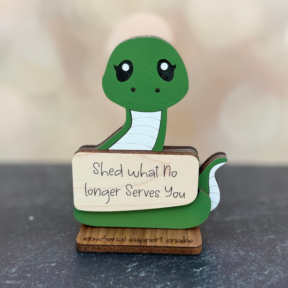 A wooden emotional support snake figurine with a green head and body, white underbelly, and a light wood sign that reads 'Shed what No longer Serves You'. The base is engraved with 'emotional support snake'.