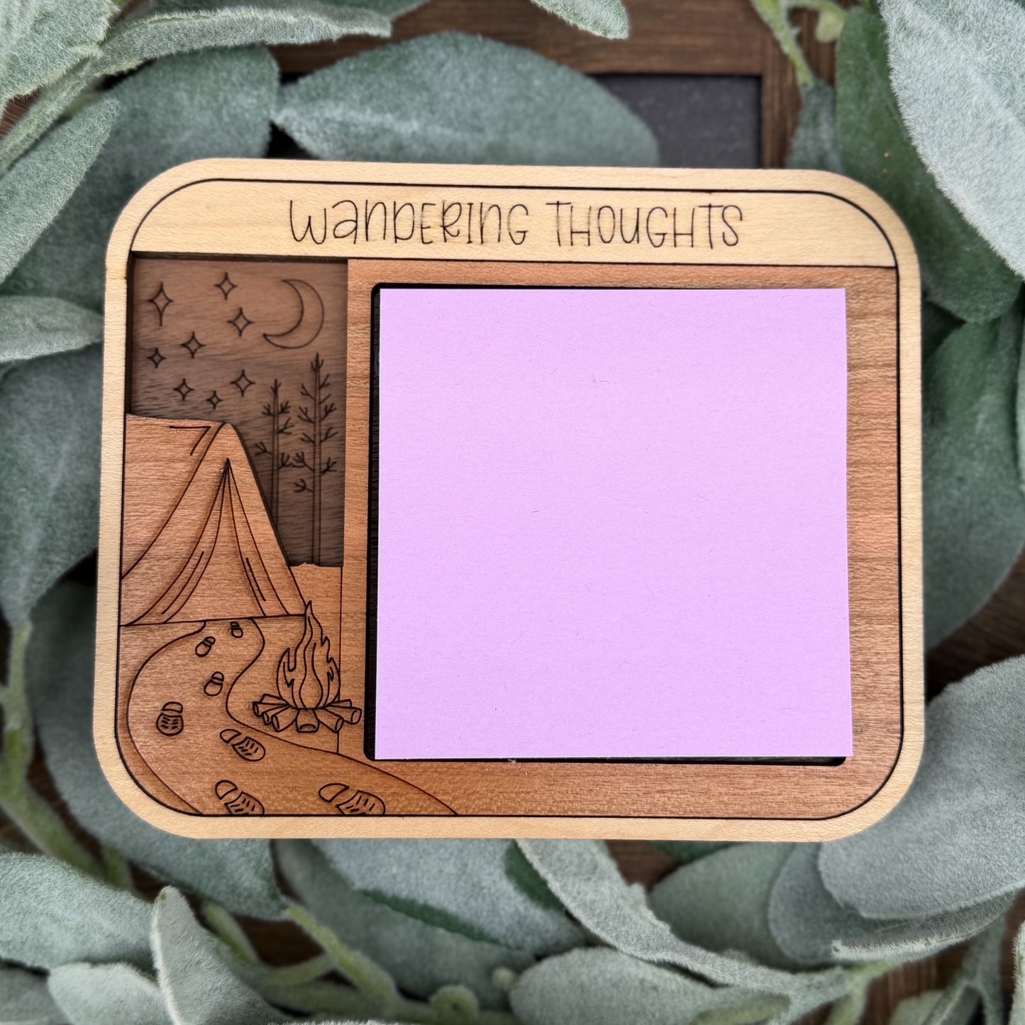 Wooden square with 'Wandering Thoughts' text, campfire, and tent design on a purple background, surrounded by green leaves.