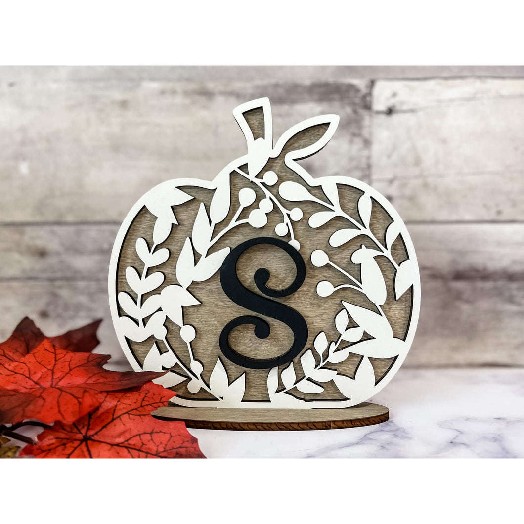 Wooden pumpkin décor with leafy cutout design and a black monogram letter “S,” displayed with red fall leaves on a rustic background.