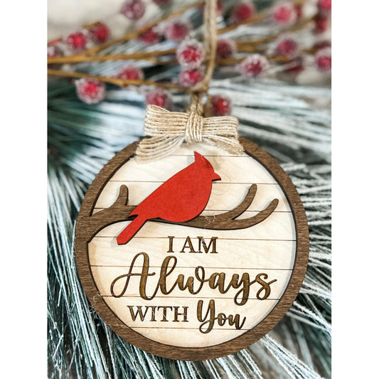 Round wooden ornament with red cardinal on branch and engraved text “I Am Always With You,” tied with rustic twine and bow on holiday greenery.