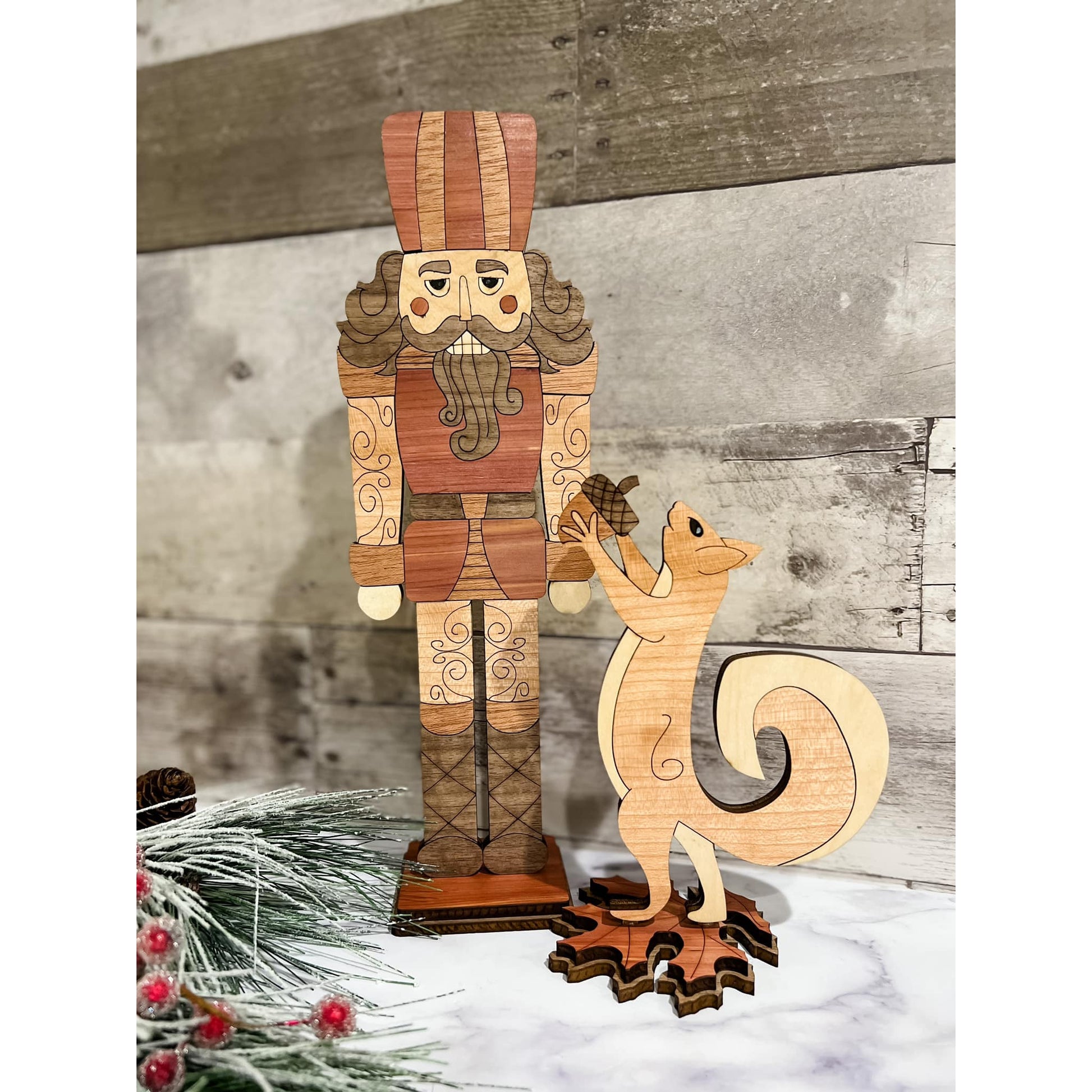 A wooden nutcracker and squirrel figurine set, with the nutcracker standing taller than the squirrel. They are placed against a wooden background with holiday decorations around.