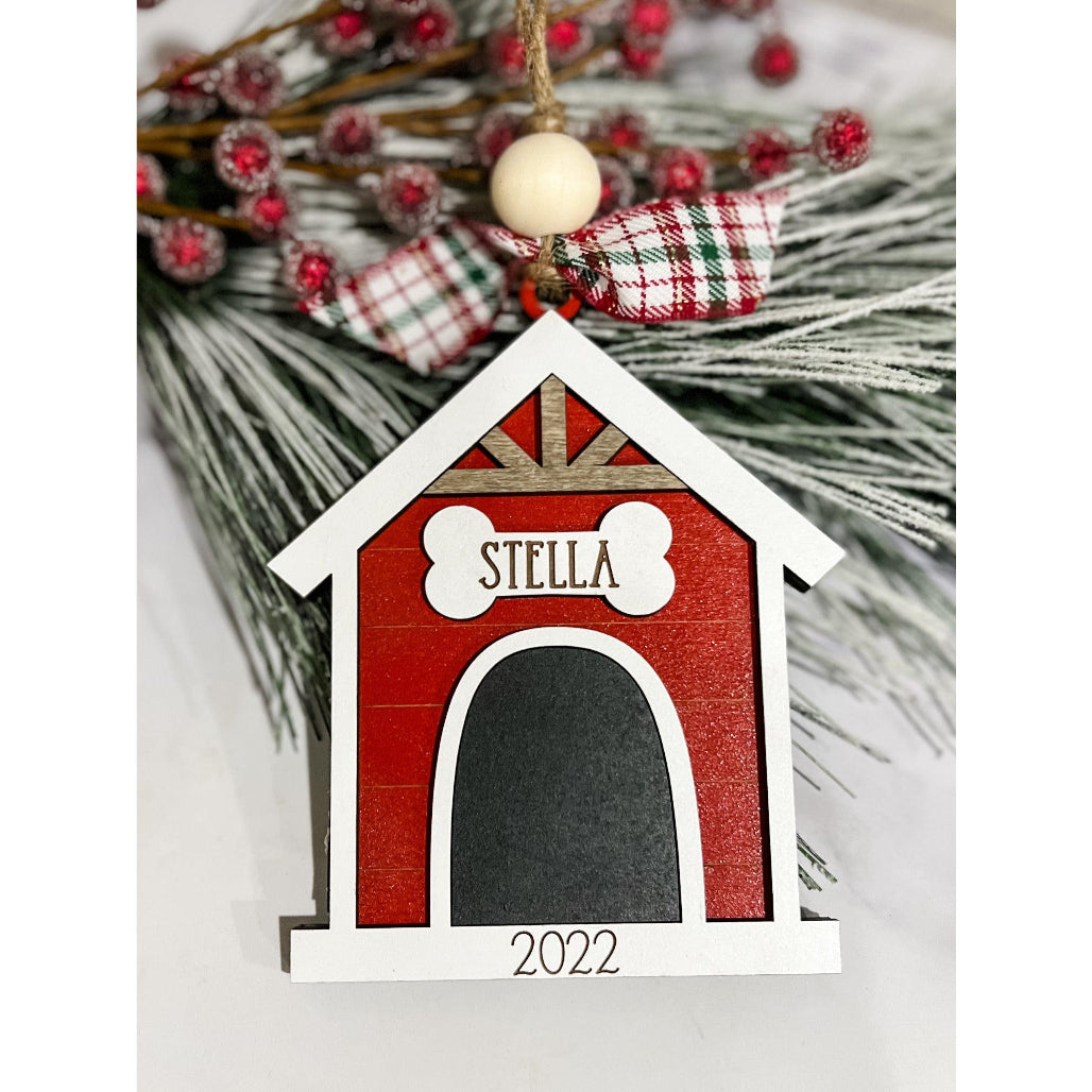 Red and white personalized doghouse Christmas ornament with engraved name “Stella,” plaid bow, and twine hanger on holiday greenery.
