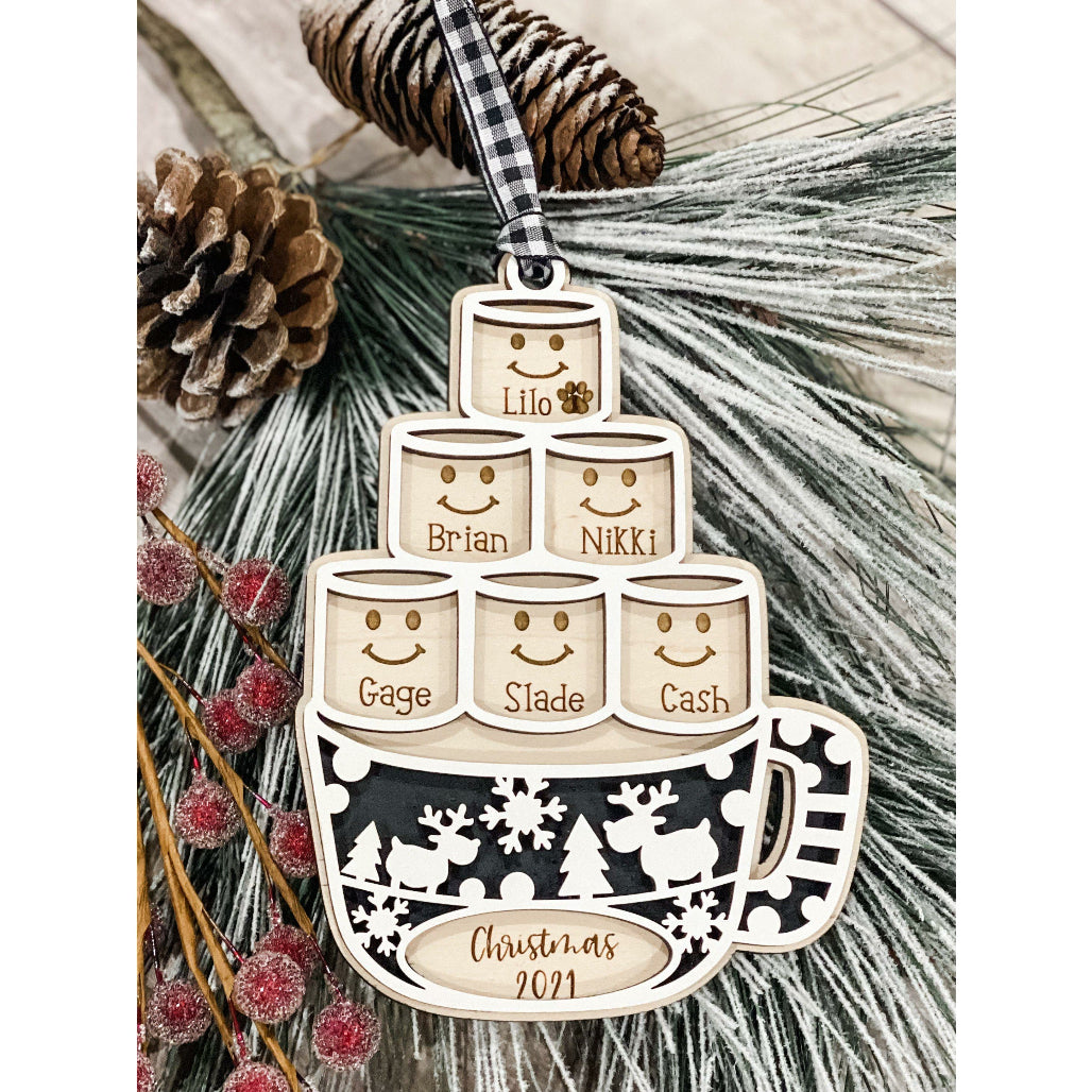 A personalized wooden marshmallow family ornament with the ability to customize with names, in the form of a stacked marshmallow shape, hanging from a string with a Christmas tree in the background.