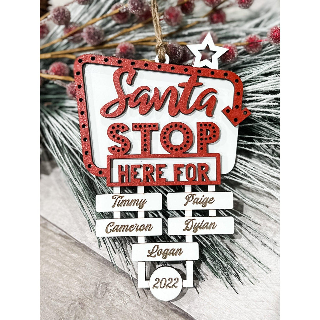 Personalized Santa Stop Here Christmas ornament with red lettering, engraved family names, and 2022 date hanging on holiday greenery.