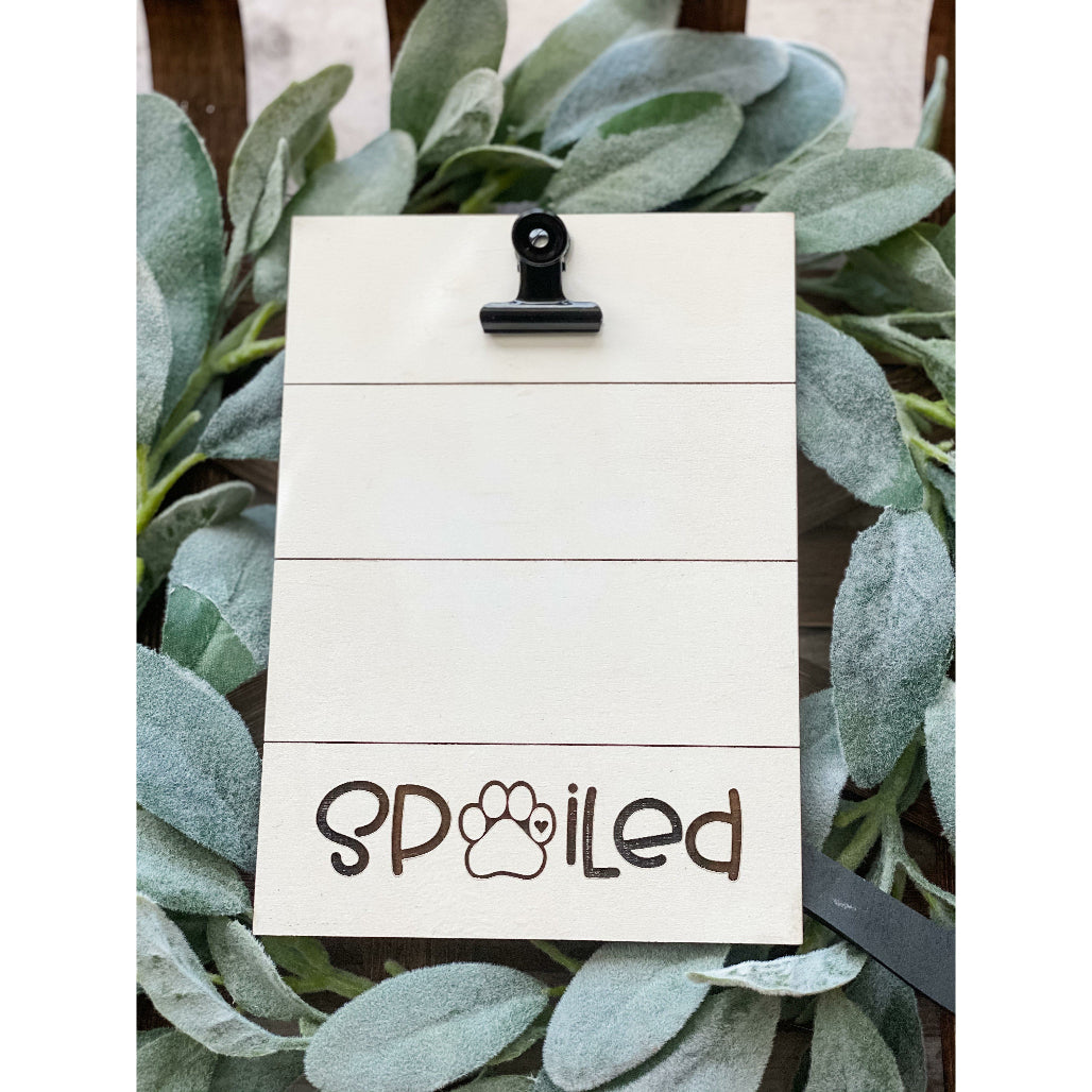 Wooden photo holder with engraved word “Spoiled” featuring paw print design and black metal clip, surrounded by greenery.
