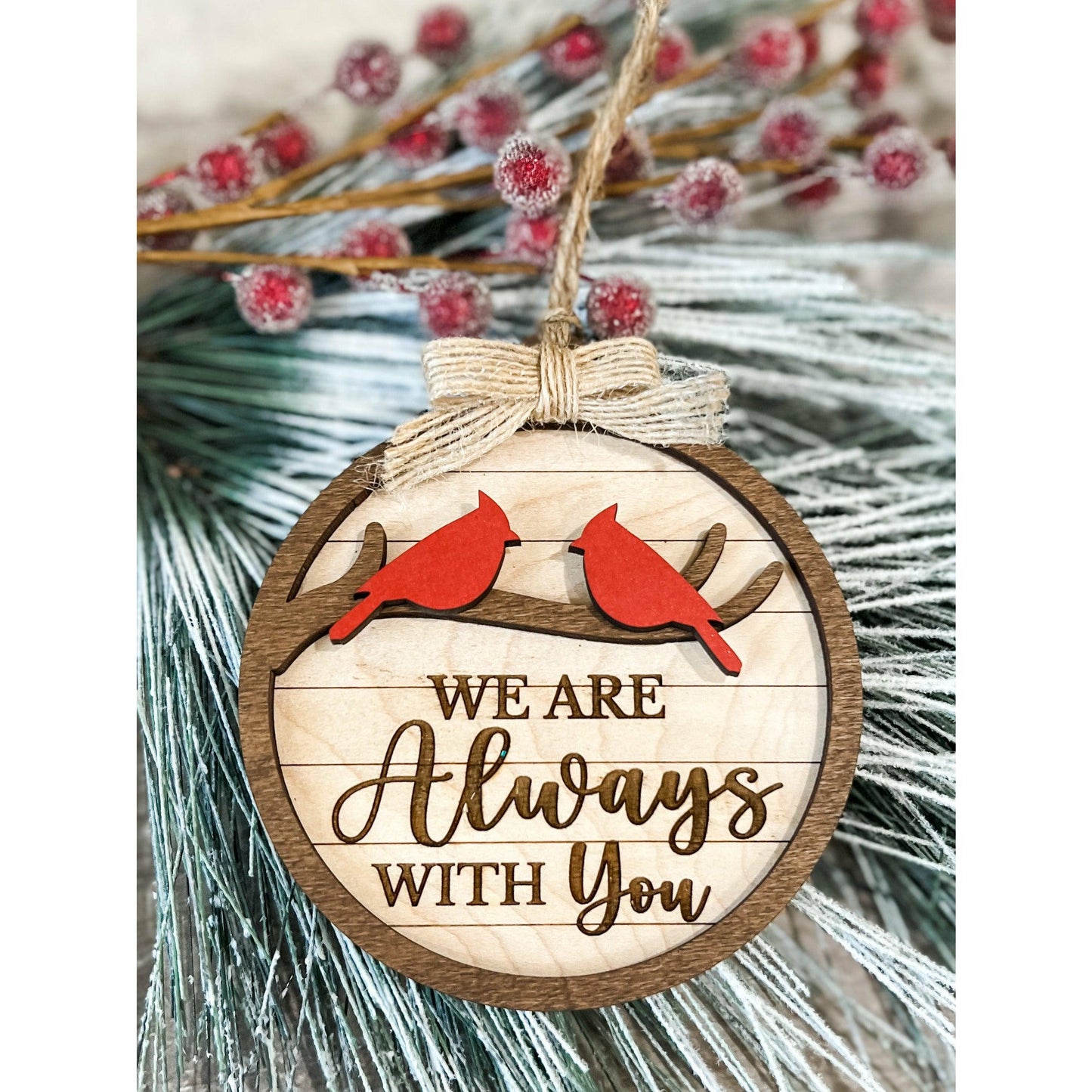 Round wooden ornament with two red cardinals on branch and engraved text “We Are Always With You,” tied with rustic twine and bow on holiday greenery.