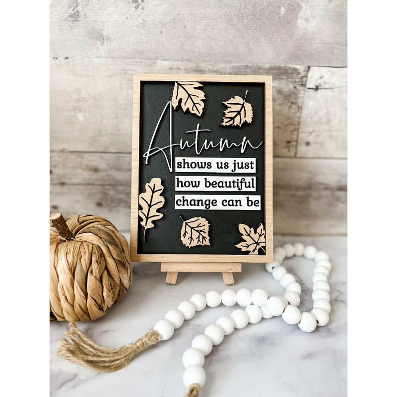 A strand of wooden beads with tassels, displayed in a clear glass vase with a leaf motif on the top, alongside a textual decoration that reads 'Autumn shows us just how beautiful change can be'.